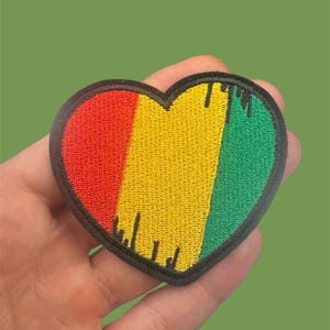 Op de afbeelding: Een hartvormige geborduurde patch met verticale strepen in rood, geel en groen. Het ontwerp bevat zwarte druppelachtige accenten langs de randen en een dikke zwarte rand die de gehele hartvorm omringt.