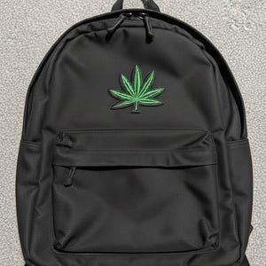 Op de afbeelding: Zwarte rugzak met een groen cannabisblad-embleem. De rugzak heeft een voorvak met rits, twee zijvakken en een handvat aan de bovenkant. Gemaakt van duurzaam materiaal.
