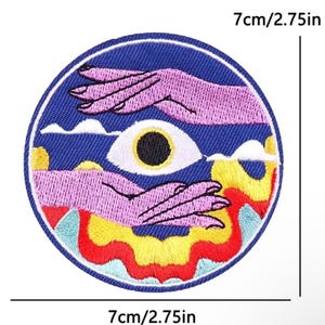 Op de afbeelding: Een ronde geborduurde patch met een diameter van 7 cm. Het ontwerp toont twee paarse handen die een centraal wit oog met een zwarte pupil omvatten, tegen een donkerblauwe achtergrond met witte wolken en gele en rode vlammen.