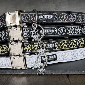 May include: A stack of four pet collars featuring a repeating pentagram pattern and a metal side-release buckle. Each collar displays a black or white fabric band with the text REBELHOUND OBEY NO MASTER and includes a dangling skull charm.