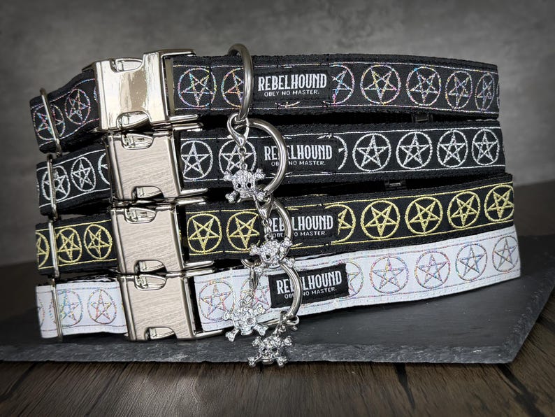 May include: A stack of four pet collars featuring a repeating pentagram pattern and a metal side-release buckle. Each collar displays a black or white fabric band with the text REBELHOUND OBEY NO MASTER and includes a dangling skull charm.