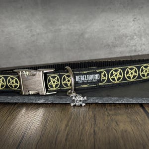 May include: A black pet collar featuring a repeating pattern of yellow pentagrams. The collar includes a silver-toned plastic side-release buckle, a metal D-ring, and a small skull-shaped charm. A label reads REBELHOUND OBEY NO MASTER.