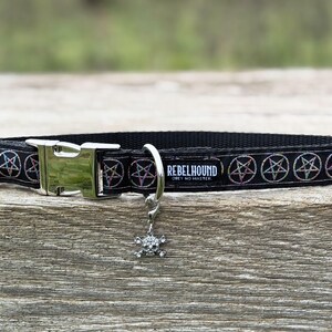 May include: A black pet collar featuring a repeating pattern of iridescent pentagrams. The collar includes a silver-toned plastic buckle, a metal D-ring, and a dangling skull charm. A black fabric tag displays the white text REBELHOUND OBEY NO MASTER.