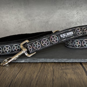 May include: A black dog leash featuring a repeating pattern of iridescent pentagrams inside circles. The leash includes a silver-toned metal swivel snap hook and a rectangular fabric label with the text REBELHOUND OBEY NO MASTER.