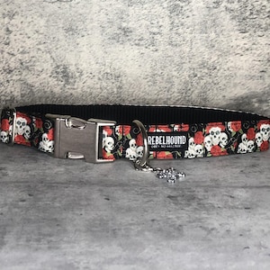 May include: Black dog collar featuring a pattern of white skulls and red roses. It has a silver metal buckle and a silver metal ring for a lead. The text "REBELHOUND OBEY NO MASTER" is printed on the collar.