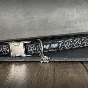 May include: A black pet collar featuring a repeating pattern of iridescent pentagrams. The collar includes a silver-toned metal side-release buckle, a D-ring, and a small skull charm. A black fabric tag displays the text REBELHOUND OBEY NO MASTER.