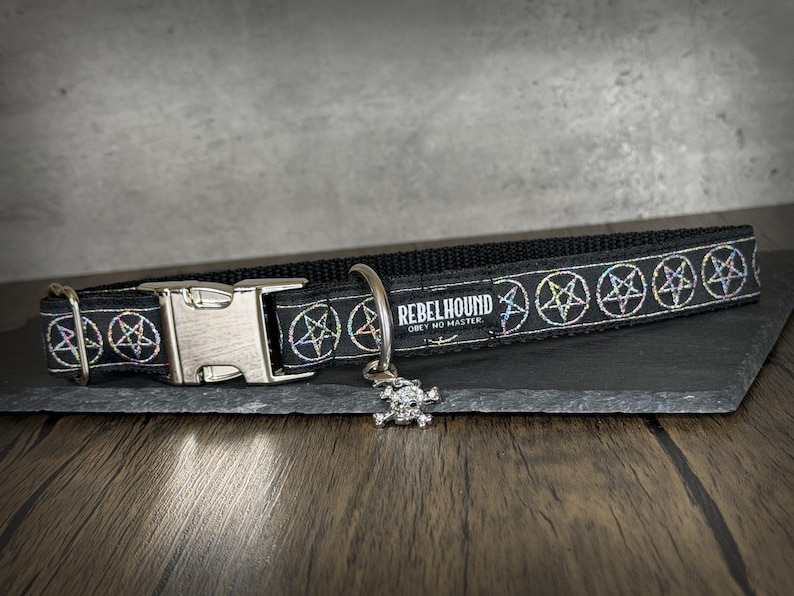 May include: A black pet collar featuring a repeating pattern of iridescent pentagrams. The collar includes a silver-toned metal side-release buckle, a D-ring, and a small skull charm. A black fabric tag displays the text REBELHOUND OBEY NO MASTER.