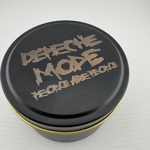 May include: A black, circular tin with a gold-coloured rim. The lid displays the words "Depeche Mode" in a gold, distressed font, with "People Are People" written underneath.