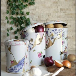 Puede incluir: Tres recipientes de almacenamiento de cocina de metal blanco con detalles florales y dorados. Los recipientes están llenos de cebollas, ajo y patatas. Los recipientes tienen tapas y asas. Una tabla de cortar de madera con cebollas está en primer plano.