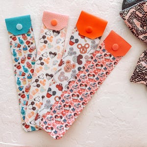 May include: Four colorful pen holders with snap closures. The patterns include cupcakes, Mickey Mouse heads, and other themed designs. The pen holders are made of a flexible material and are in various colors, including teal, pink, and orange.