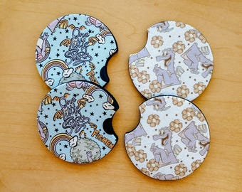 May include: Four car coasters with a whimsical design. Two coasters feature a light blue background with rainbows, stars, and text that reads "One Little Spark" and "Figment". The other two have a repeating pattern of a purple dragon and flowers.