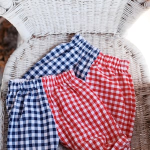 May include: Three pairs of gingham patterned shorts in red and navy blue, resting on a white wicker chair. The shorts have elastic waistbands and rolled cuffs, perfect for warm weather.