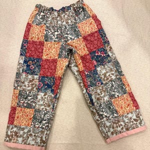 Puede incluir: Pantalones patchwork con una variedad de patrones florales y geométricos en tonos rojos, azules, beige y verdes. Los pantalones tienen un ribete rosa en la parte inferior y una cintura elástica. Una prenda única y colorida.
