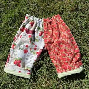 May include: Shorts with a strawberry design. One side is covered in a pattern of red strawberries, the other has a white background with strawberry illustrations and text. The shorts have a green and white striped trim.