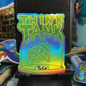 May include: A holographic sticker with the words "THINK TANK" in green dripping letters above a green brain inside a glass dome. The sticker has a rainbow gradient background and the text "Tx Cx" at the bottom.