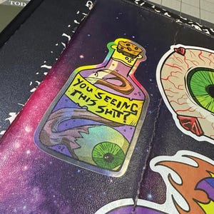 May include: A holographic sticker featuring a cartoon potion bottle with a cork stopper. The bottle contains a green eyeball and the text "YOU SEEING THIS SHIT?". The sticker is on a notebook with other stickers.