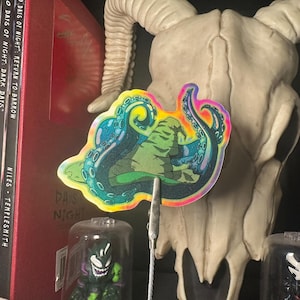 May include: A colorful sticker featuring an octopus design with green, blue, and yellow accents. The sticker is held by a metal clip in front of a skull and other collectibles. The sticker has the words "DAYS NIGHT" on it.