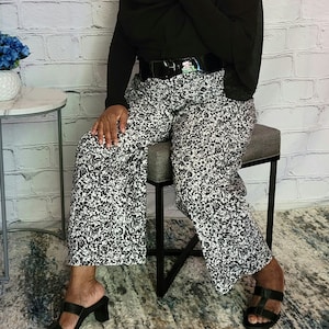 May include: A person wearing a white wide-brimmed hat with a shell trim, a black long-sleeved shirt, black belt, and black and white patterned pants. The person is sitting on a gray stool with black legs.