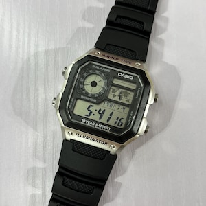 May include: A Casio digital watch with a silver-tone rectangular case and a black textured resin strap. The watch face features a world map display, a circular analogue-style sub-dial, and a digital time readout showing 5:41:16 with 10 YEAR BATTERY text.