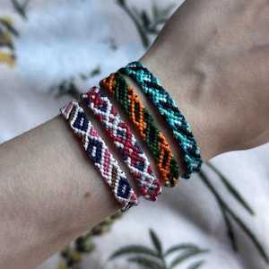 May include: Four woven friendship bracelets featuring intricate geometric patterns. The first bracelet uses white, navy, and pink thread. The second uses white, red, and navy. The third uses orange, green, and black. The fourth uses teal and black.