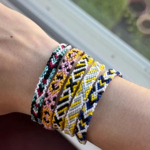 May include: A collection of colorful friendship bracelets with intricate woven patterns. The bracelets feature a variety of colors, including red, black, pink, yellow, white, and blue. The bracelets are displayed on a person's arm.