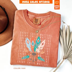 May include: A yam-colored t-shirt with the text "EVERY CHILD MATTERS" repeated around a graphic of colorful feathers. The shirt has a crew neck and short sleeves. A gold necklace and a brown hat are visible.