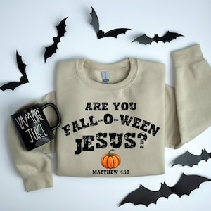 May include: A beige sweatshirt with the text "ARE YOU FALL-O-WEEN JESUS?" and a pumpkin graphic. A black mug with "VAMPIRE JUICE" is on the left. Black bat cutouts are scattered around the sweatshirt, creating a Halloween theme.