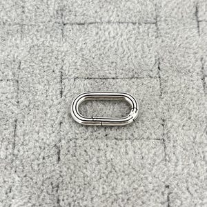 Könnte beinhalten: Ein silberfarbener, ovaler Verschluss mit einem Scharnierdesign für einen sicheren Verschluss. Der Verschluss befindet sich vor einem strukturierten grauen Hintergrund, der seinen metallischen Glanz und seine komplizierten Details hervorhebt. Ideal für die Schmuckherstellung oder das Basteln.