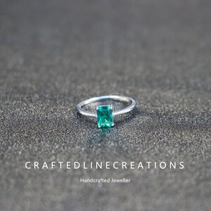 Peut inclure: Une bague en argent ornée d'une pierre précieuse rectangulaire vert émeraude. La bague est posée sur une surface sombre et texturée. Le texte "CRAFTEDLINECREATIONS" et "Handcrafted Jeweller" sont en bas.
