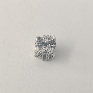 May include: A silver charm in the shape of a gift box with a bow. The bow and box are embellished with clear, sparkling stones. The charm is made of 925 silver.