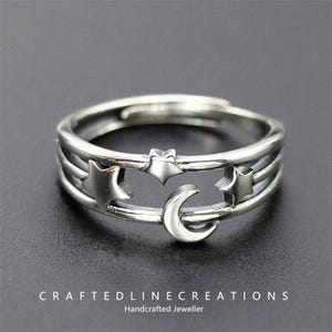 Puede incluir: Un anillo plateado con un diseño celestial con estrellas y una luna creciente. El anillo tiene una banda ajustable y está elaborado con detalles intrincados. Es una pieza de joyería artesanal. El texto "CRAFTEDLINECREATIONS Handcrafted Jeweller" está en la parte inferior.