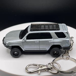 May include: Silver SUV keyring with black wheels, a roof rack, and a spare tyre. The keyring has a silver clasp and a large silver ring. The vehicle is a miniature model.