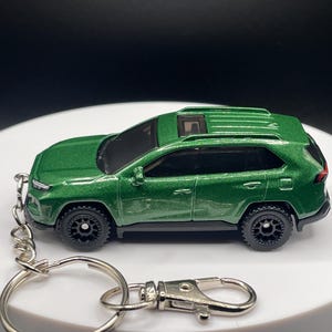 May include: A green die-cast SUV keyring with black wheels and a silver ring. The miniature vehicle has a detailed design, including a sunroof and realistic features. The keyring is a compact accessory.