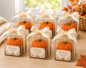 May include: Six clear square boxes each containing an orange pumpkin-shaped candle with a green stem. Each box is topped with a cream-colored satin ribbon bow and features a label that reads Thank you FOR CELEBRATING WITH US with a small pumpkin icon.