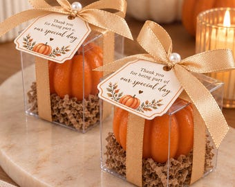 Puede incluir: Dos cajas de regalo cuadradas transparentes con objetos naranjas en forma de calabaza sobre papel marrón arrugado. Cada caja tiene una cinta dorada, una perla blanca y una etiqueta que dice: Thank you for being part of our special day.