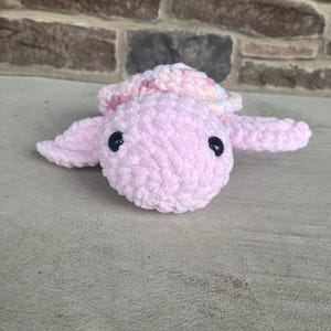 May include: A handmade, plush turtle in soft pink yarn. The turtle has a textured body, a multi-colored shell, and black safety eyes. The turtle's flippers are extended, and it is lying on a neutral surface.