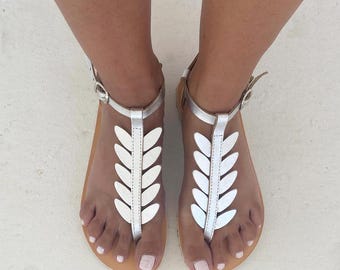 May include: A pair of silver sandals featuring a t-strap design adorned with a series of metallic leaf-shaped cutouts running down the center. The sandals include a thin ankle strap with a small silver buckle closure and a flat tan sole.