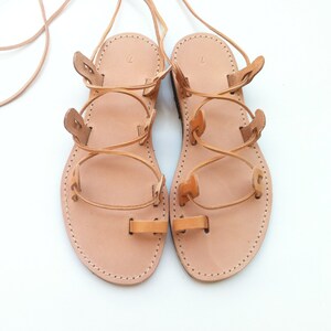 Op de afbeelding: Een paar beige leren sandalen met een teenlus, meerdere zijbandjes en lange veters om rond de enkel te wikkelen. Het voetbed is gestempeld met het cijfer 7 en de randen zijn afgewerkt met zichtbare witte stiksels langs de zool.