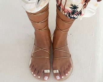 May include: Metallic gold gladiator sandals featuring thin straps that wrap around the ankles and a wide, flat band across the big toe. The sandals have a simple, open design with a flat sole, providing a minimalist look that highlights the gold finish.