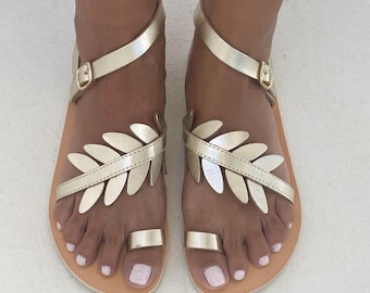 May include: A pair of gold sandals featuring a leaf-shaped design across the top, a toe ring, and an adjustable ankle strap with a metal buckle. The sandals have a flat tan sole and a metallic finish, providing a secure fit around the foot.