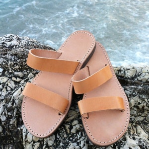 A pair of tan leather sandals with two straps. The sandals are on a rocky surface.