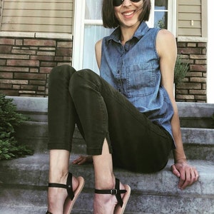 May include: A woman wearing a denim shirt and dark green pants sits on a set of concrete steps. She is wearing black sandals with a leopard print strap.