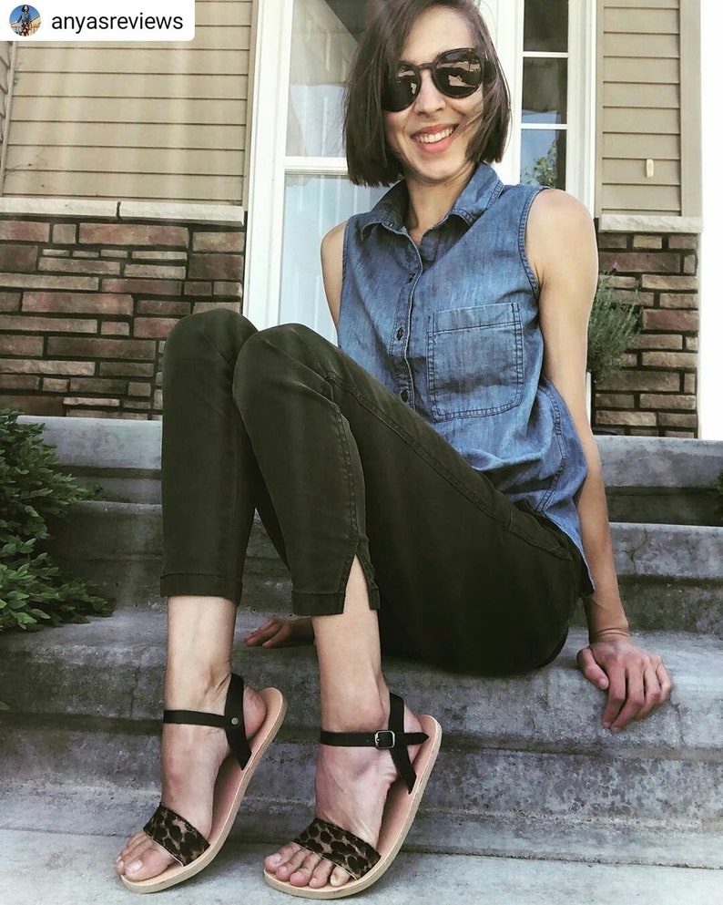 May include: A woman wearing a denim shirt and dark green pants sits on a set of concrete steps. She is wearing black sandals with a leopard print strap.