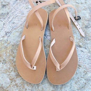 May include: A pair of tan leather sandals with a simple design. The sandals have a single strap that wraps around the ankle and a toe loop. The sandals are size 38.