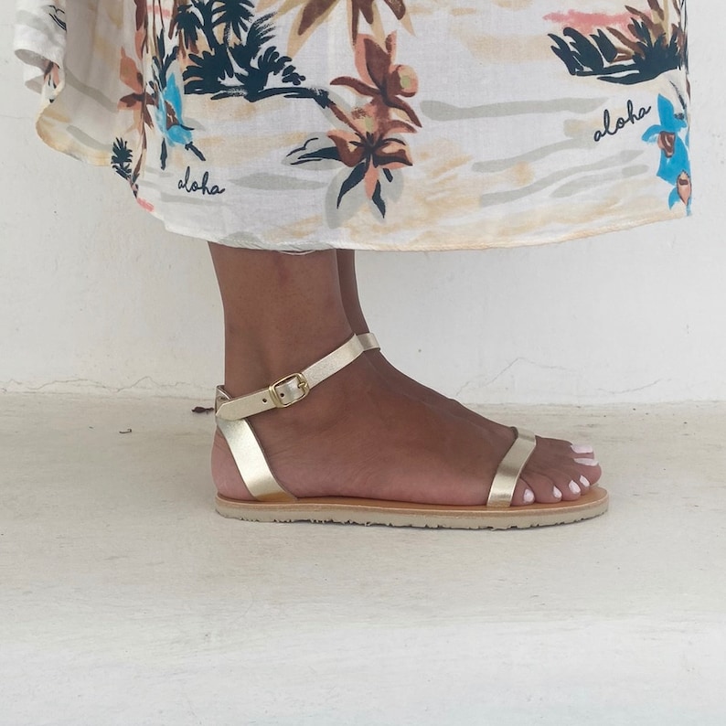 May include: A metallic gold flat sandal featuring a thin ankle strap with a gold-toned metal buckle and a single horizontal toe strap. The sandal has a flat, light-colored sole and is worn beneath a white skirt with a tropical floral print.