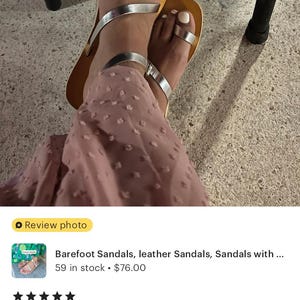 May include: A pair of flat sandals featuring metallic silver straps, including a toe loop design and an ankle strap, set against a tan-colored footbed. The sandals are worn with a dusty rose-colored garment with a textured, dotted pattern.