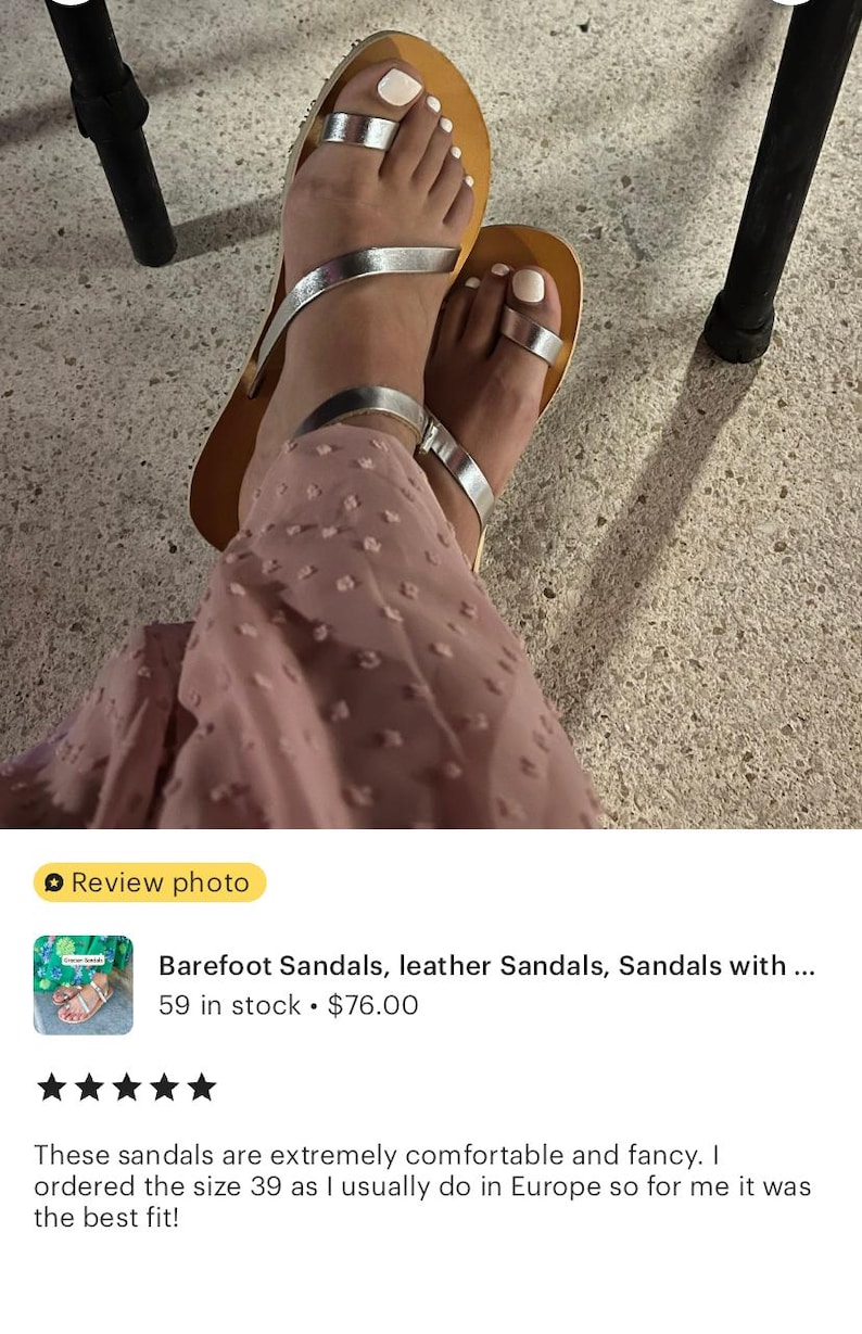 May include: A pair of flat sandals featuring metallic silver straps, including a toe loop design and an ankle strap, set against a tan-colored footbed. The sandals are worn with a dusty rose-colored garment with a textured, dotted pattern.