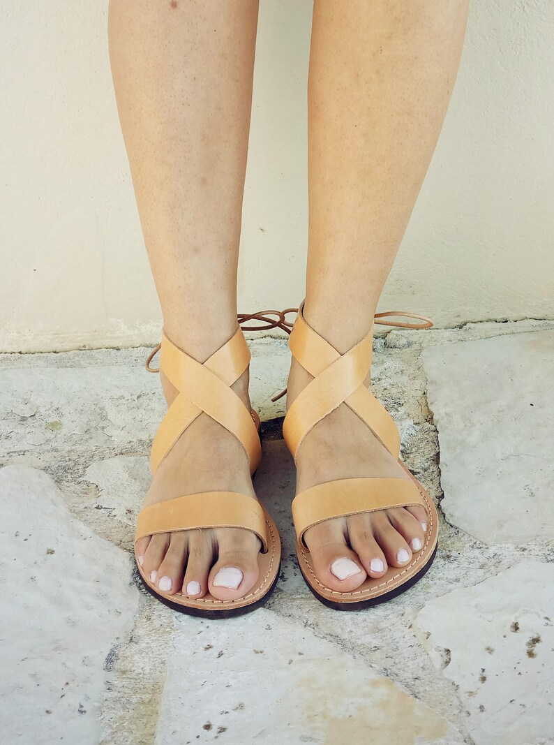 May include: A pair of light tan leather sandals featuring a single horizontal strap across the toes and two crisscrossing straps that wrap around the ankle, secured with thin leather ties at the back of the heel and a dark brown sole.
