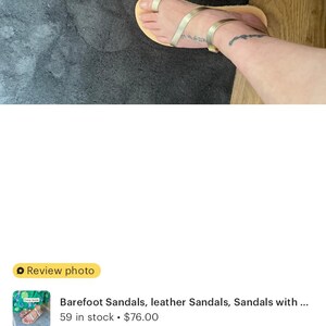May include: A pair of gold-colored sandals featuring a thin strap design that wraps around the big toe and across the midfoot, secured with a slim ankle strap and a small metallic buckle, set on a flat, light-colored sole.