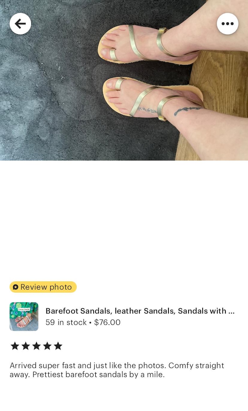 May include: A pair of gold-colored sandals featuring a thin strap design that wraps around the big toe and across the midfoot, secured with a slim ankle strap and a small metallic buckle, set on a flat, light-colored sole.